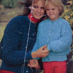 May include: A woman and a child wearing matching blue and teal knitted cardigans with a textured, bobbly design. The woman is wearing a red scarf and the child is wearing red trousers.