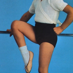 May include: A young girl wearing a white knitted top with a wrap-style neckline and black shorts. She is standing on her toes with one leg raised and holding onto a bar. She is wearing white socks and brown ballet shoes.