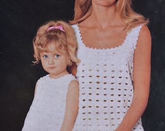 May include: Two people wearing white crocheted dresses with pink and orange flower accents. The dress on the left has a shorter hemline and the dress on the right has a longer hemline.