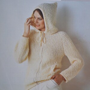Pode incluir: Camisola com capuz de malha creme com fecho de cordão e fecho de correr frontal. A camisola tem mangas compridas e um padrão de malha texturizada. O capuz é pontudo no topo. A pessoa está a usar calças brancas.