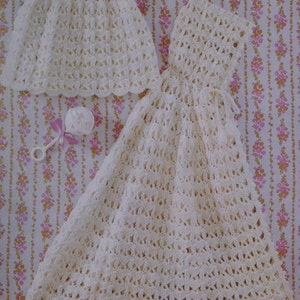 Peut inclure: Deux robes de bébé en crochet, l'une à manches courtes et l'autre longue, toutes deux en blanc. Les robes sont disposées sur un tissu à motifs floraux.