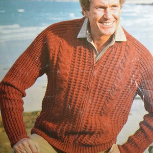 May include: A red knitted cardigan with a zipper closure. The cardigan has a cable knit pattern and is worn by a person standing on a beach.