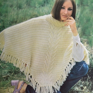 Könnte beinhalten: Ein weißer gestrickter Poncho mit einem Zopfmuster und Fransenbesatz. Der Poncho wird von einer Person mit braunen Haaren getragen und sitzt auf einer grasbewachsenen Fläche.