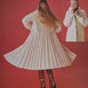 Op de afbeelding: Een lange witte gebreide jas met een kabelbreipatroon. De jas heeft een lange, vloeiende rok en een hoge kraag. Het model draagt ​​bruine laarzen.