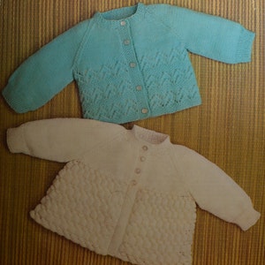 May include: Two knitted baby cardigans, one in a light blue color with a lacy pattern and the other in a cream color with a textured pattern.