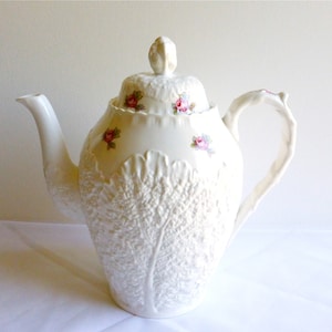 Może przedstawiać: Biały porcelanowy czajnik z wzorem liścia kapusty i różowymi akcentami kwiatowymi. Czajnik ma pokrywkę z gałką w kształcie kwiatu.