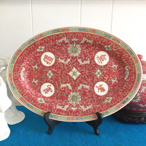 May include: A red oval ceramic serving platter with a decorative floral pattern and Chinese characters. The platter is on a black stand.