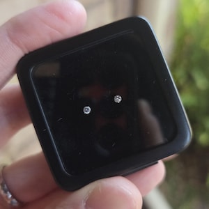 May include: A pair of small, round, clear faceted gemstones displayed inside a square black plastic storage case with a transparent lid. The stones are positioned centrally against the dark interior, highlighting their reflective surfaces.