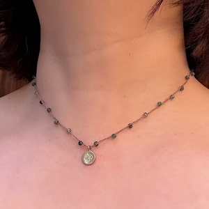 May include: A delicate gold necklace with a small round pendant featuring an engraved Om symbol. The necklace is made with a thin chain and small green beads.