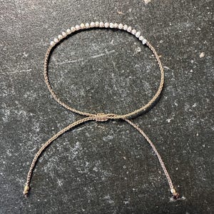 May include: A delicate bracelet featuring a row of small, white beads strung along a thin, woven, gold-colored cord. The bracelet has a sliding knot closure with two dangling cord ends. The jewelry is displayed on a dark, textured surface.