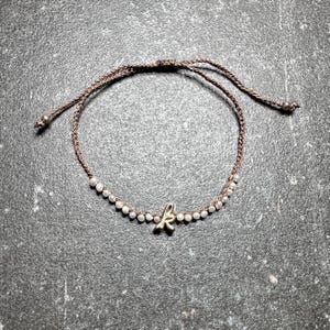 May include: A brown macrame bracelet with small white beads and a gold-colored letter "k" charm. The bracelet is adjustable with sliding knots. The beads are evenly spaced around the bracelet.