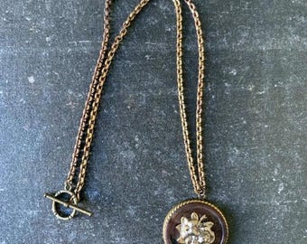 May include: A gold chain necklace with a round pendant featuring a floral design. The pendant is made of dark brown metal with gold accents.