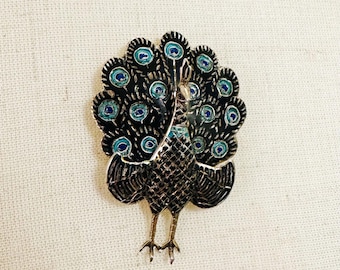 May include: A black and blue enamel peacock brooch with a detailed design. The peacock's tail feathers are spread out in a fan shape, and the bird's head is turned to the side.