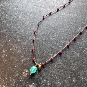 May include: Delicate gold necklace featuring small red gemstones and a larger, oval-shaped, light blue-green gemstone pendant. A small gold-colored teardrop charm hangs next to the pendant. The necklace is displayed on a dark, textured surface.