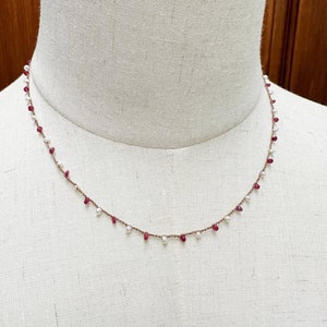 Può includere: Una collana delicata con perle bianche e pietre preziose rosse alternate. La collana è su uno spago marrone ed è esposta su un manichino bianco.