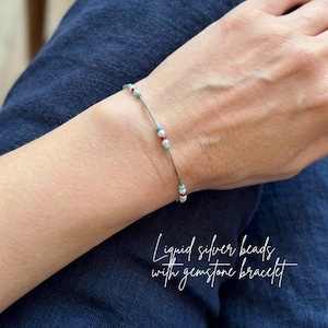 May include: A delicate bracelet featuring liquid silver beads and gemstone accents. The bracelet has a thin silver chain with small, colorful beads in shades of red, blue, and white. The text "Liquid silver beads with gemstone bracelet" is written in cursive.