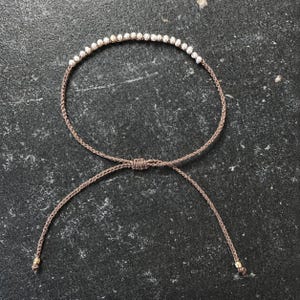 Könnte beinhalten: Ein zartes Armband mit einer Reihe kleiner, weißer, runder Perlen, die auf eine dünne, braune, geflochtene Kordel gefädelt sind. Das Armband verfügt über einen verstellbaren Schiebeknoten und zwei lose Kordelenden, die mit einer kleinen Goldperle abschließen.