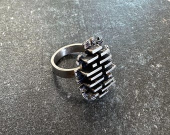 May include: A silver ring with a unique, textured design. The ring features a series of stacked, rectangular shapes, creating a geometric and modern aesthetic. The band is smooth and simple, contrasting with the detailed centerpiece.