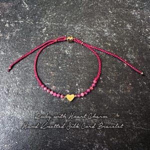 May include: A ruby bracelet with a gold heart charm. The adjustable bracelet is made with a knotted silk cord. The text on the image reads "Ruby with Heart Charm Hand Knotted Silk Cord Bracelet."