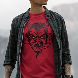 May include: A man wearing a red t-shirt with a black and white illustration of a devil's face with horns and a cigar. He is wearing a green and black plaid shirt over the t-shirt.