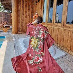Puede incluir: Una bata larga hasta el suelo de color rosa empolvado con un intrincado bordado floral multicolor en la espalda. El diseño muestra grandes peonías amarillas y rosas, flores blancas más pequeñas y follaje verde, con mangas largas y anchas y un dobladillo fluido.