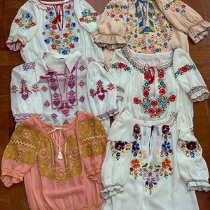 Puede incluir: Una colección de seis blusas con diversos patrones de bordado floral y geométrico. Las prendas incluyen telas blancas, color melocotón y beige con detalles de hilos coloridos, escotes con lazos y mangas fruncidas con puños de volantes.