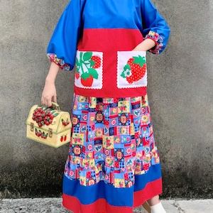Könnte beinhalten: Ein blau-rotes Langarmshirt mit zwei weißen Taschen mit Erdbeermotiven, kombiniert mit einem langen, bunten Patchworkrock mit Blumen- und Geometriemustern sowie einer beigen Handtasche, die mit dreidimensionalen roten Erdbeeren verziert ist.
