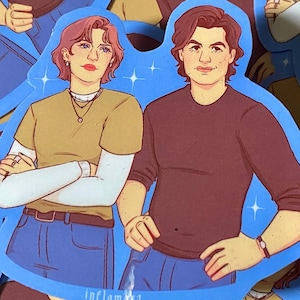 May include: Sticker sheet featuring multiple stickers with illustrations of two people. The stickers have a blue outline with star accents. The illustrations depict a person with red hair and a person with dark hair, both wearing casual clothing.