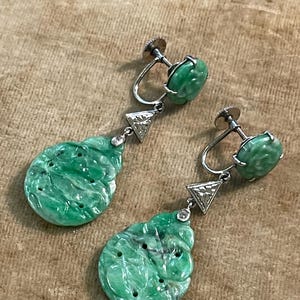 May include: A pair of green jade drop earrings with a carved design. The earrings are set in silver and feature a small diamond accent.