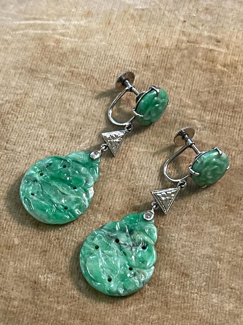 May include: A pair of green jade drop earrings with a carved design. The earrings are set in silver and feature a small diamond accent.