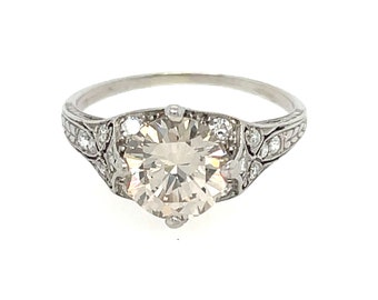 May include: A platinum ring featuring a large, round-cut diamond as the centerpiece. The ring has smaller diamonds set on the sides and intricate filigree detailing along the band. The ring's design is classic and elegant.