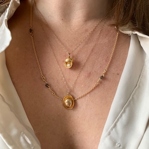 May include: Three gold necklaces with different pendants. The top necklace has a small, round pendant with a pearl in the center. The middle necklace has a simple, round pendant. The bottom necklace has a chain with small, dark beads and a larger, round pendant.