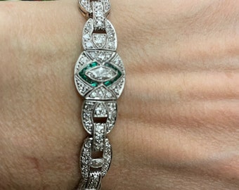 May include: A silver bracelet with a central oval-shaped design featuring a large emerald-cut gemstone surrounded by smaller diamonds. The bracelet is made of a chain of linked ovals, each set with diamonds.
