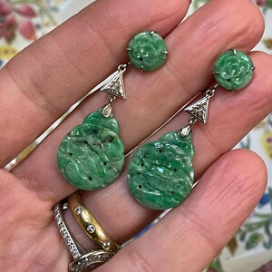 May include: A pair of green jade drop earrings with diamond accents. The earrings feature a round jade stone with a carved floral design, followed by a triangular diamond and a larger, carved jade drop.