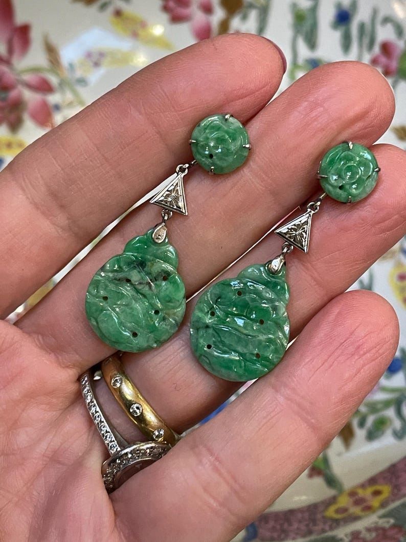 May include: A pair of green jade drop earrings with diamond accents. The earrings feature a round jade stone with a carved floral design, followed by a triangular diamond and a larger, carved jade drop.