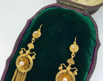 May include: A pair of gold drop earrings with pearl accents, displayed in a velvet-lined box. The earrings feature a delicate floral design and a dangling chain with small beads.