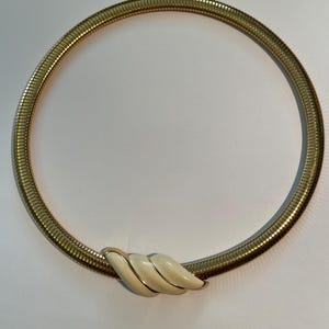 May include: A gold-toned, coiled necklace with a cream-colored, seashell-shaped clasp. The necklace has a flexible, tubular design. The clasp is a stylized, three-part design with gold trim. The necklace is a choker style.