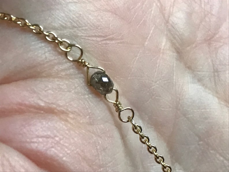 May include: A delicate gold chain necklace with a single, dark grey diamond bead.