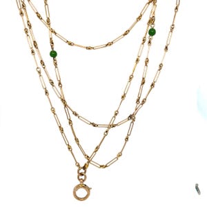 Puede incluir: Un collar de cadena en tono dorado con eslabones rectangulares alargados intercalados con segmentos trenzados. El collar incluye dos pequeñas cuentas redondas de color verde vibrante a lo largo de la cadena y termina en un cierre de resorte circular.
