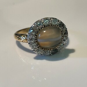 Puede incluir: Un anillo de oro con una piedra de ágata gris y blanca engastada en un halo de pequeños diamantes.