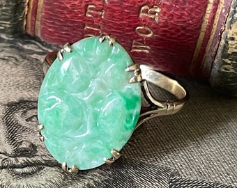 May include: A silver ring with a carved green jade stone. The stone is oval shaped and has a detailed floral design. The ring is set on a book with the words "Literary Works" visible.