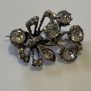Puede incluir: Un broche de plata con un diseño floral y brillantes de cristal transparentes. El broche tiene la forma de un abanico con un grupo de brillantes en la parte superior.