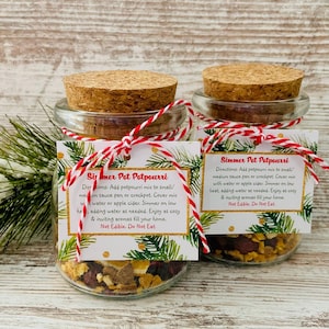 May include: Two glass jars filled with potpourri, each sealed with a cork lid and tied with red and white twine. The jars have labels with instructions. The potpourri includes dried fruits and spices, creating a cozy, inviting aroma for the home.