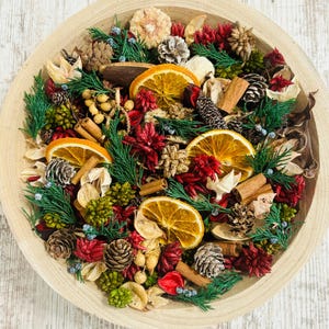 May include: A wooden bowl filled with a decorative potpourri arrangement. The arrangement includes dried orange slices, pine cones, cinnamon sticks, and sprigs of green and red foliage. The bowl is light brown and sits on a light wood surface.
