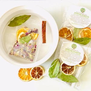 May include: Two clear plastic bags filled with dried orange slices, bay leaves, and a cinnamon stick. The bags have a white label with the text "Sweet N Salty Bath Drops Spring Scents" and a small green tree logo. A white bowl with water and a similar bag of dried ingredients is in the background.
