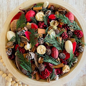 May include: A wooden bowl filled with a festive potpourri arrangement. The mix includes pine cones, red and white dried flowers, cinnamon sticks, and green sprigs. A string of wooden beads and a tassel are visible in the foreground.