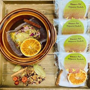 May include: Four clear plastic bags with labels that read "Simmer Pot Potpourri Sachets". Each bag contains a sachet with dried fruit, flowers, and spices. The sachets are designed to be simmered in water to release a pleasant aroma. The bags are arranged on a wooden surface with a brown ceramic bowl containing a similar sachet, a cinnamon stick, and a dried orange slice.