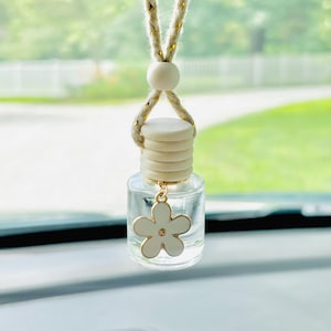 May include: Clear glass car air freshener with a white flower charm. The air freshener hangs from a beige and gold rope. The background is a blurred view of a green lawn and trees.