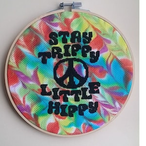 May include: A round, colorful, tie-dye fabric embroidery hoop with the text "Stay Trippy Little Hippy" and a peace sign.
