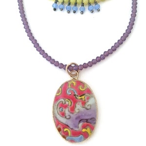 May include: A layered necklace with a purple and green beaded chain and a pendant featuring a pink and purple oval-shaped ceramic tile with a floral design.